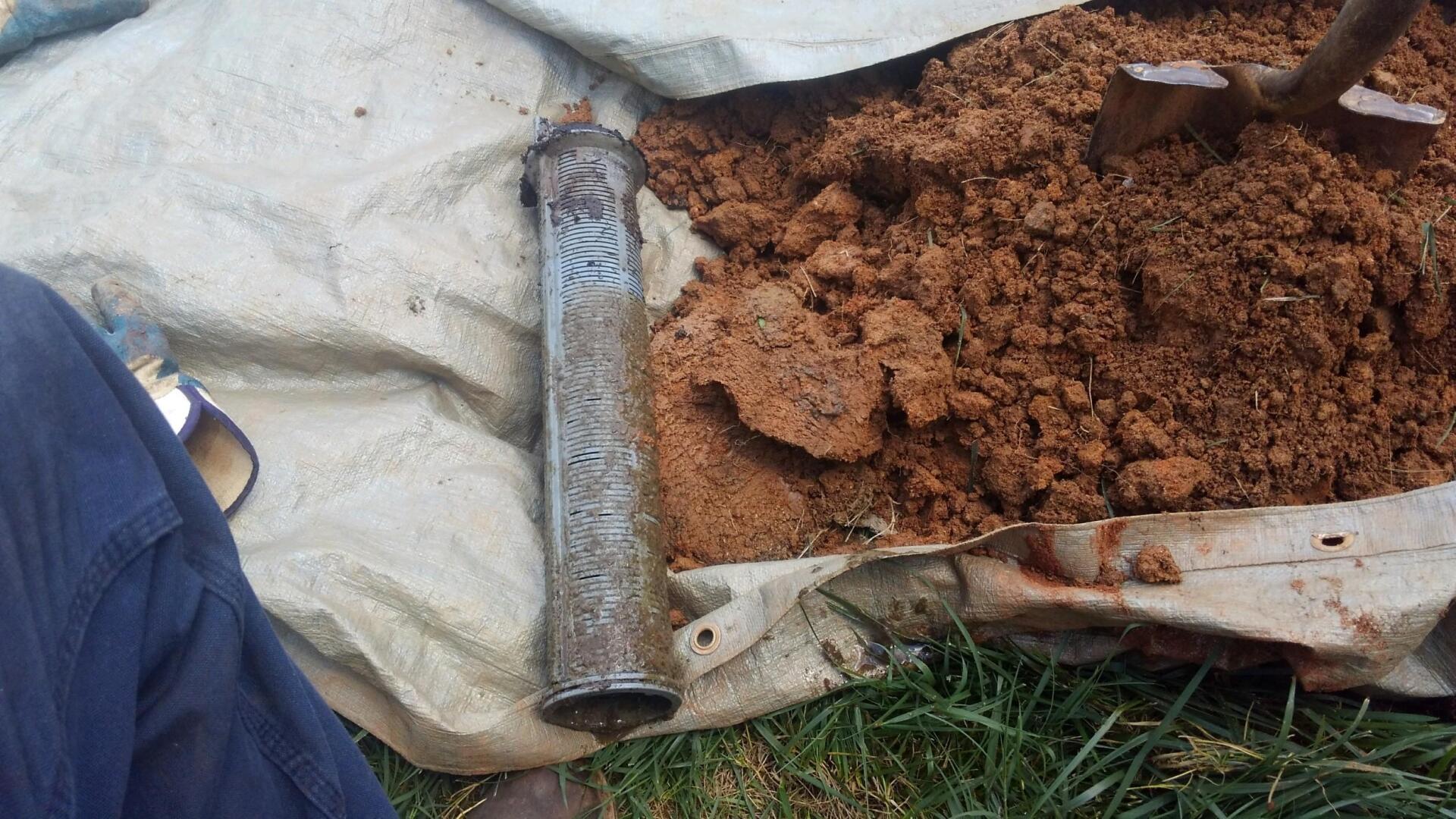 A rusty metal pipe lies on a white tarp next to a pile of brown dirt, with a person's blue jeans visible.