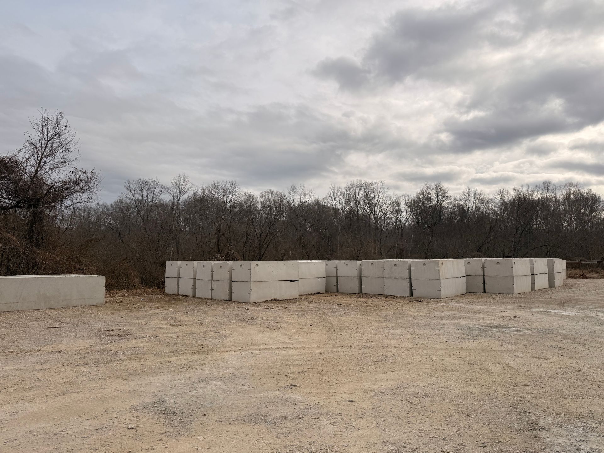 Gray concrete blocks stacked outdoors on gravel, with trees in background under cloudy sky.