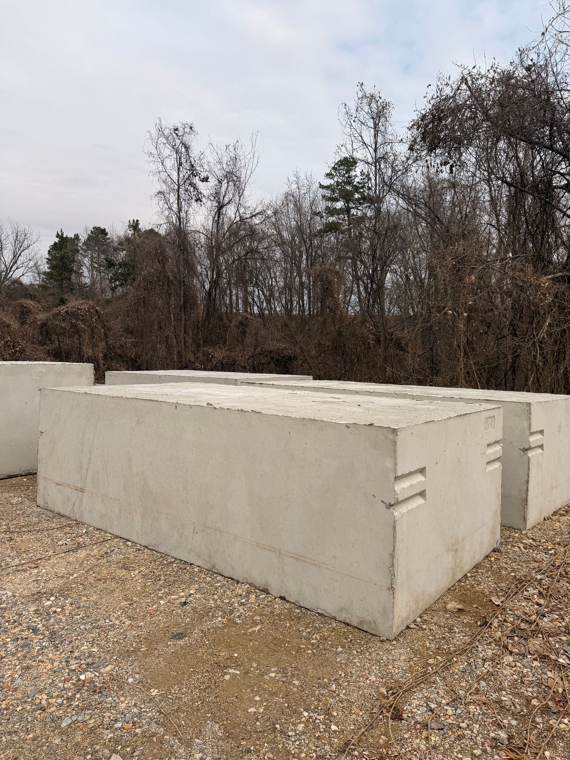 Concrete rectangular blocks stacked on gravel, with trees in the background under a cloudy sky.