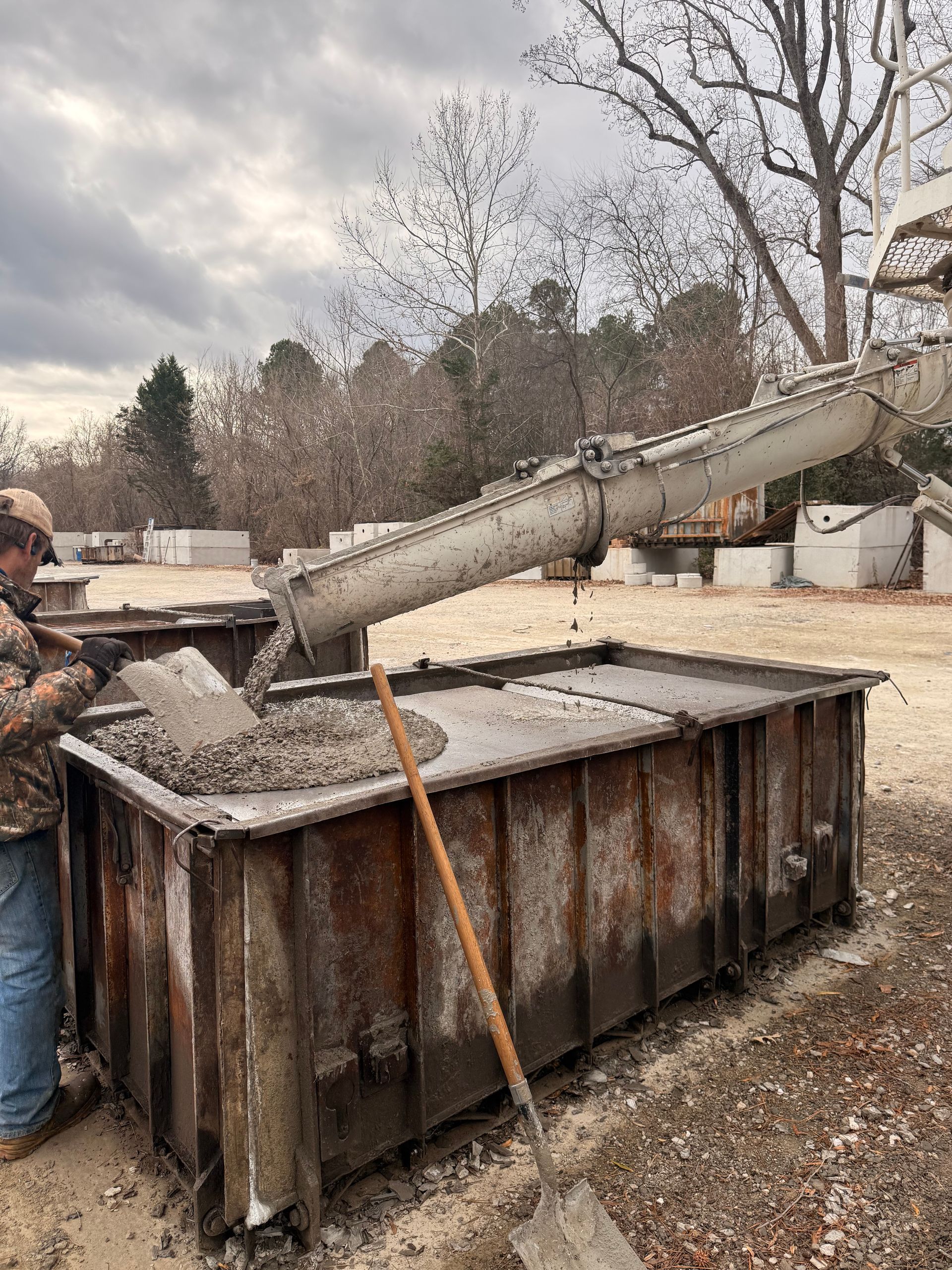 Construction worker pouring concrete into a large metal container with a chute, outdoor setting.