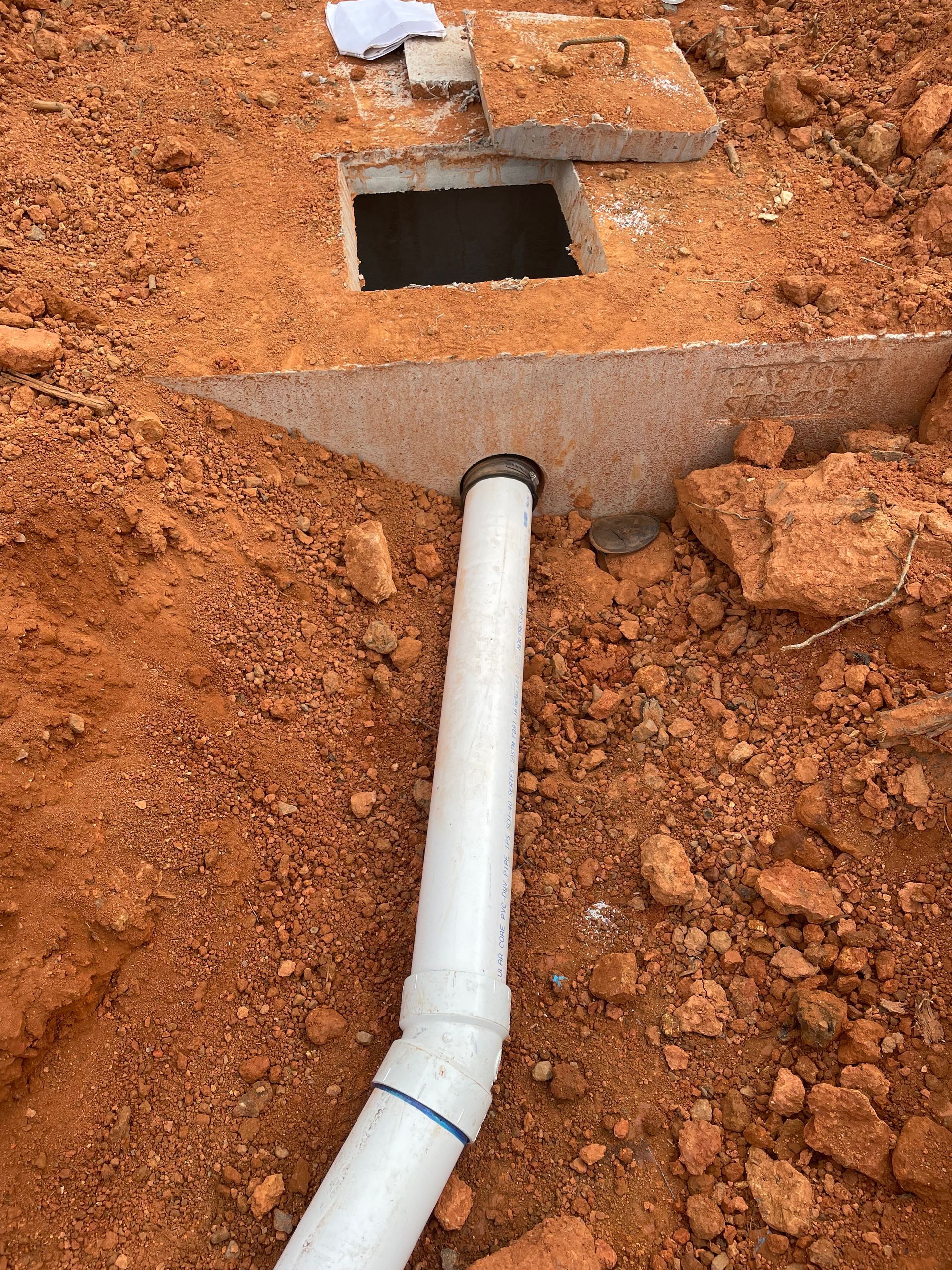 White pipe connected to an open concrete box in reddish-brown dirt.