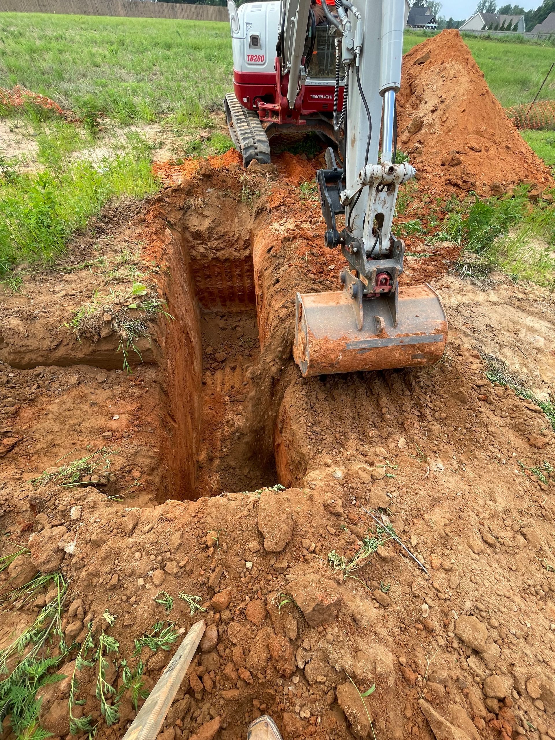 Mini excavator digging a trench in reddish-brown soil on a grassy field, with a pile of dirt in the background.