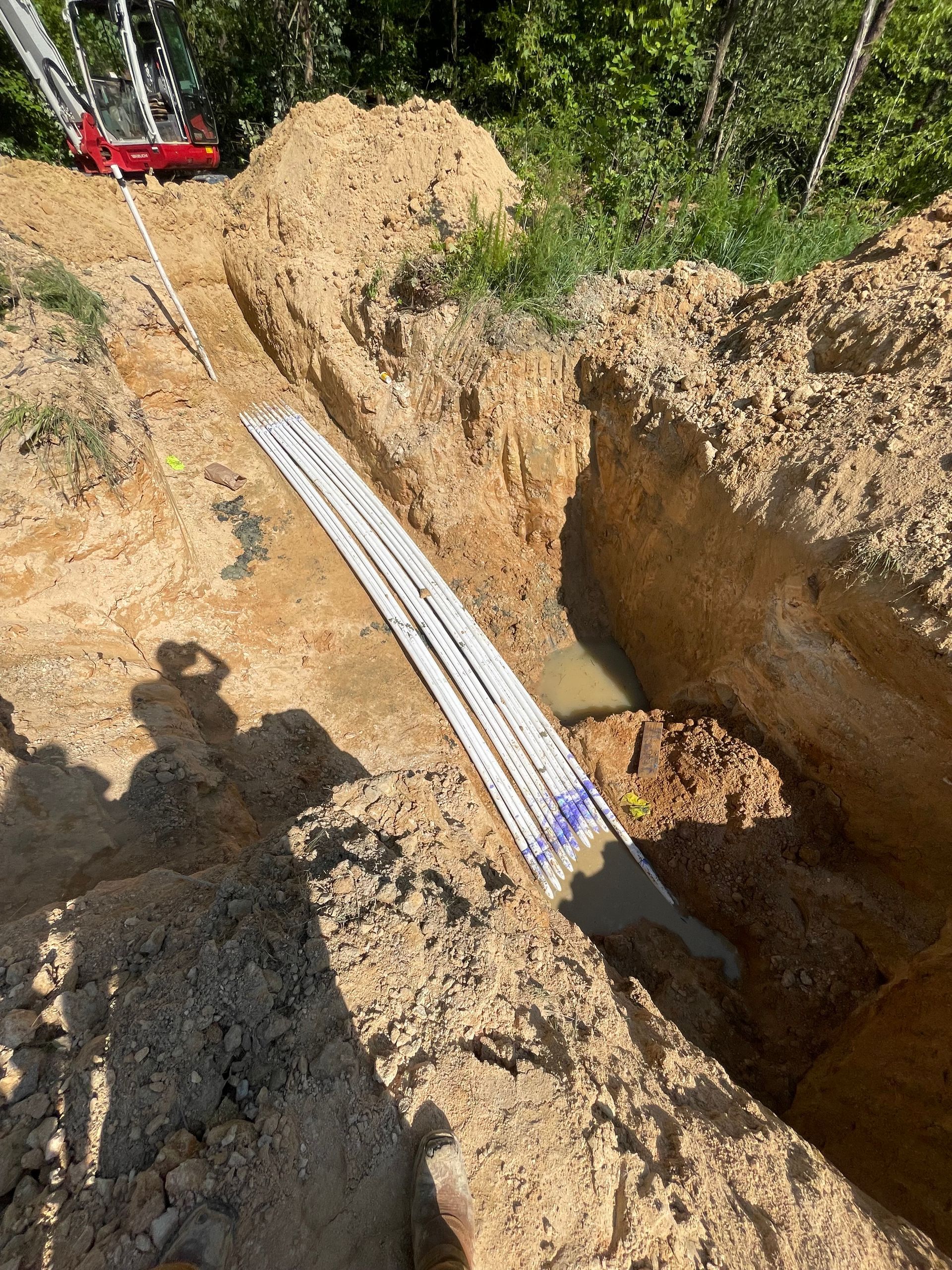Trench with a white and blue measuring tape, filled with water and dirt, with a red machine visible in the background.