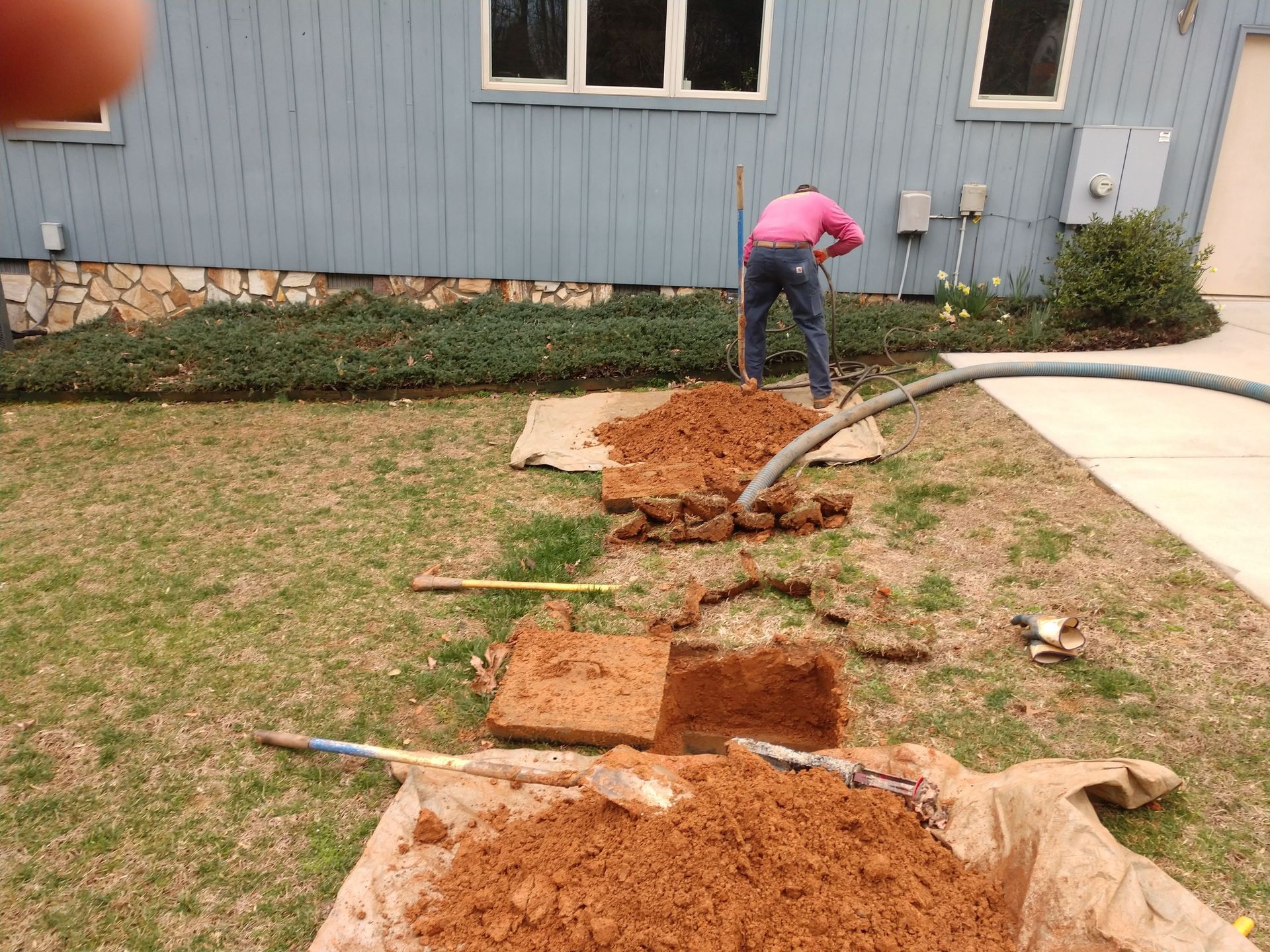 Person digging trench in lawn near house; soil piles on cardboard; hose nearby.