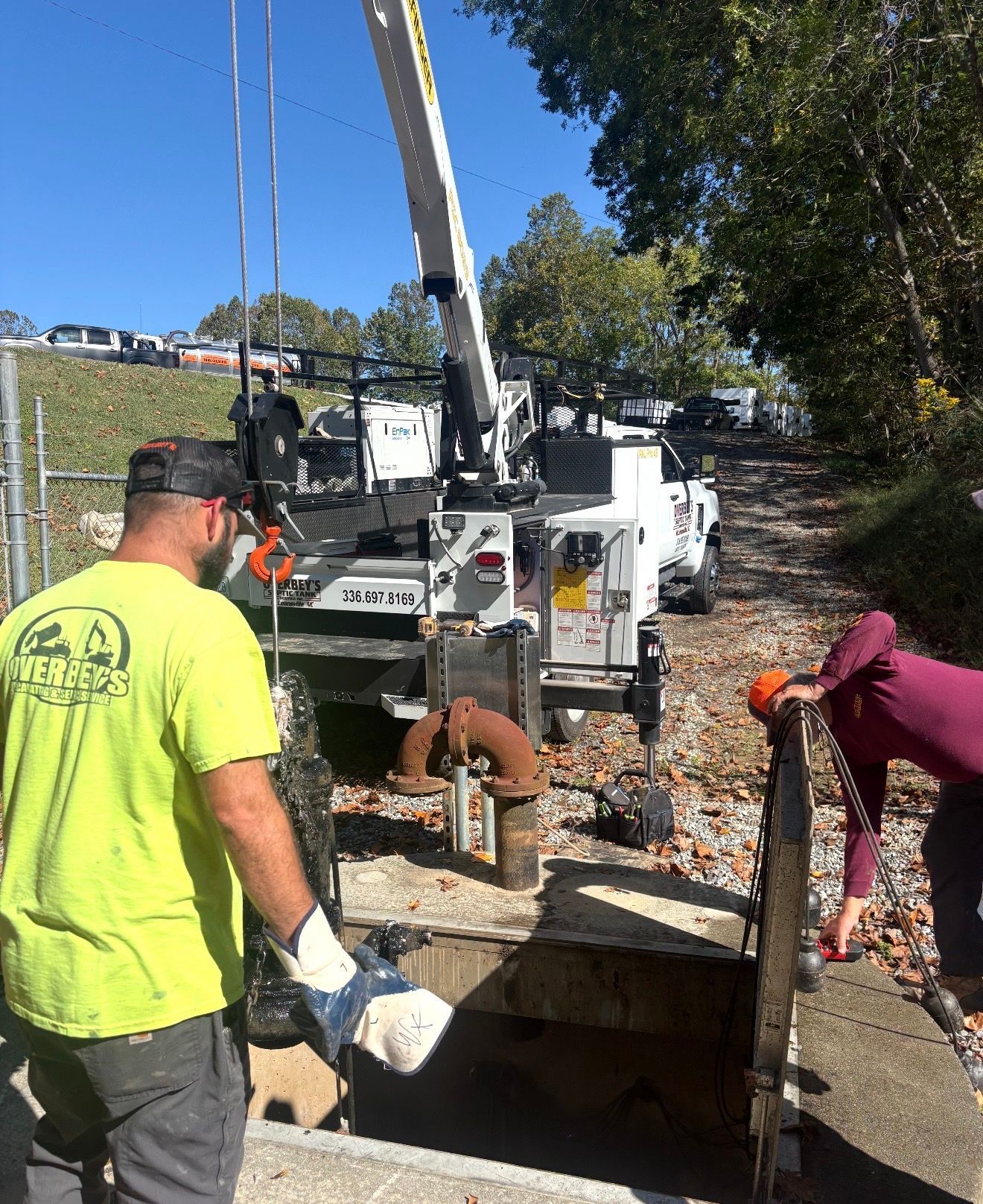 Two people in work gear operate a crane to lift a heavy metal cover from a ground-level opening on a gravel path.