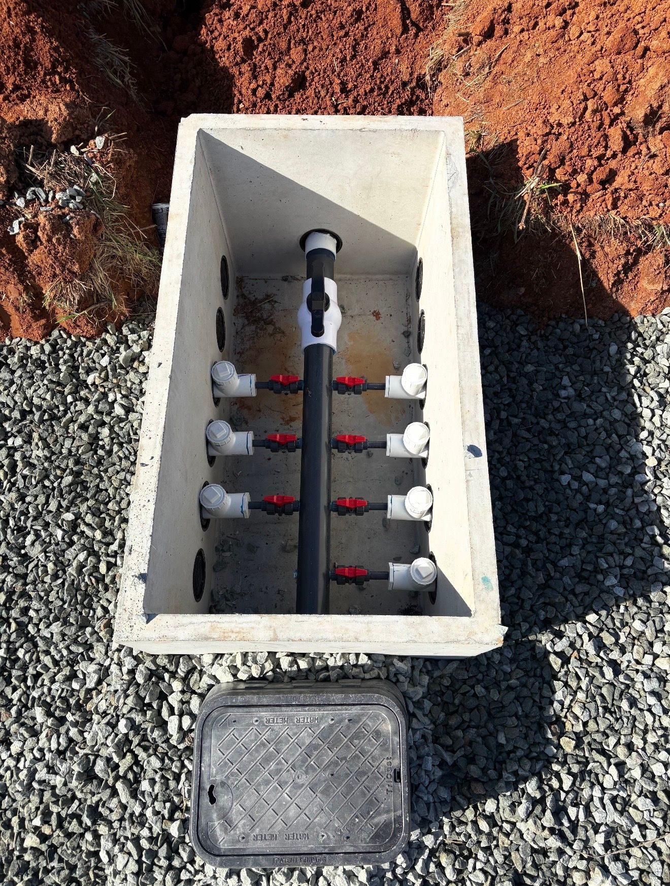 An open irrigation valve box set in gravel containing a black supply manifold with multiple red-handled valves.