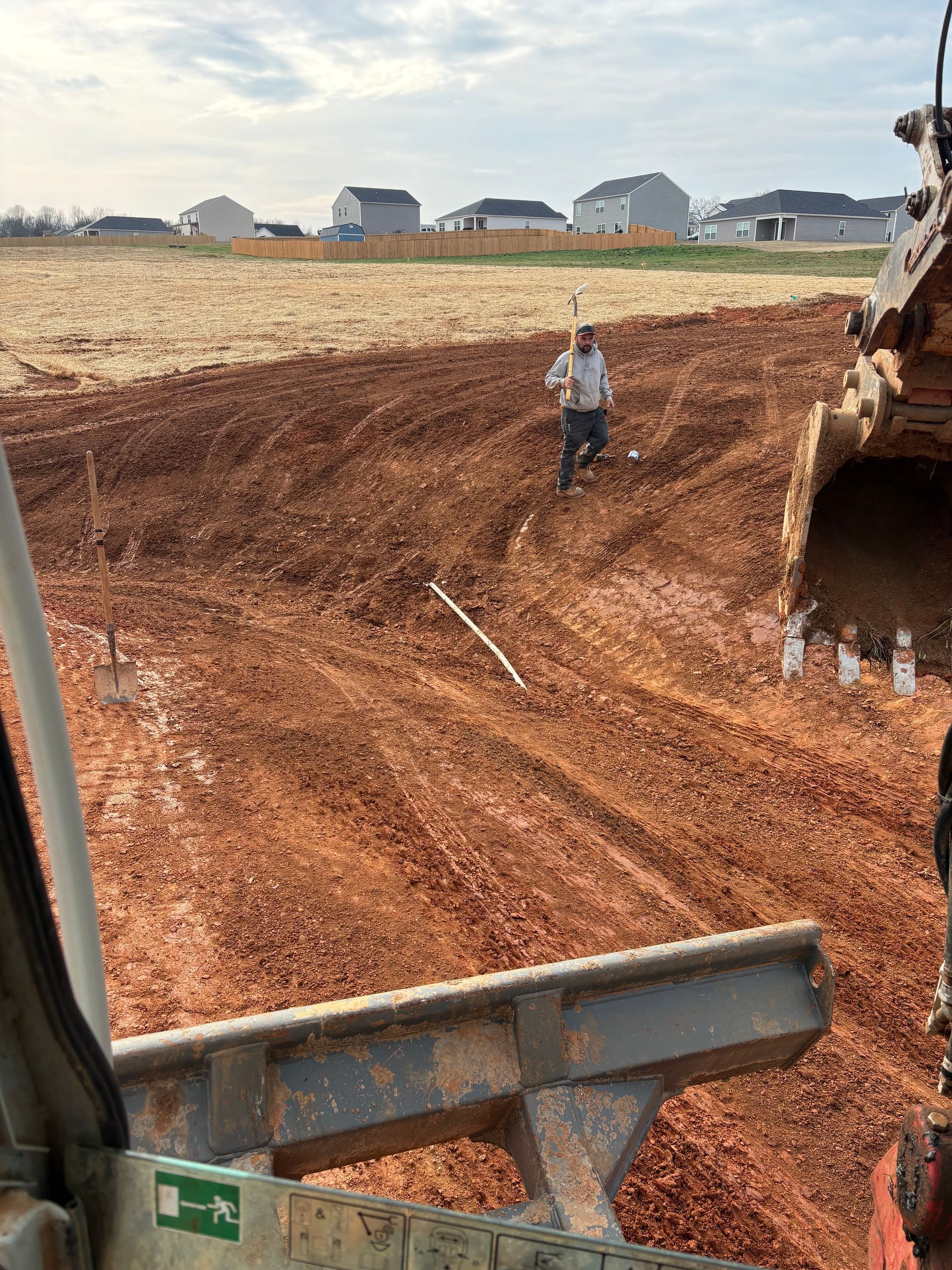 A person stands in a muddy, excavated construction site next to heavy machinery under a cloudy sky.