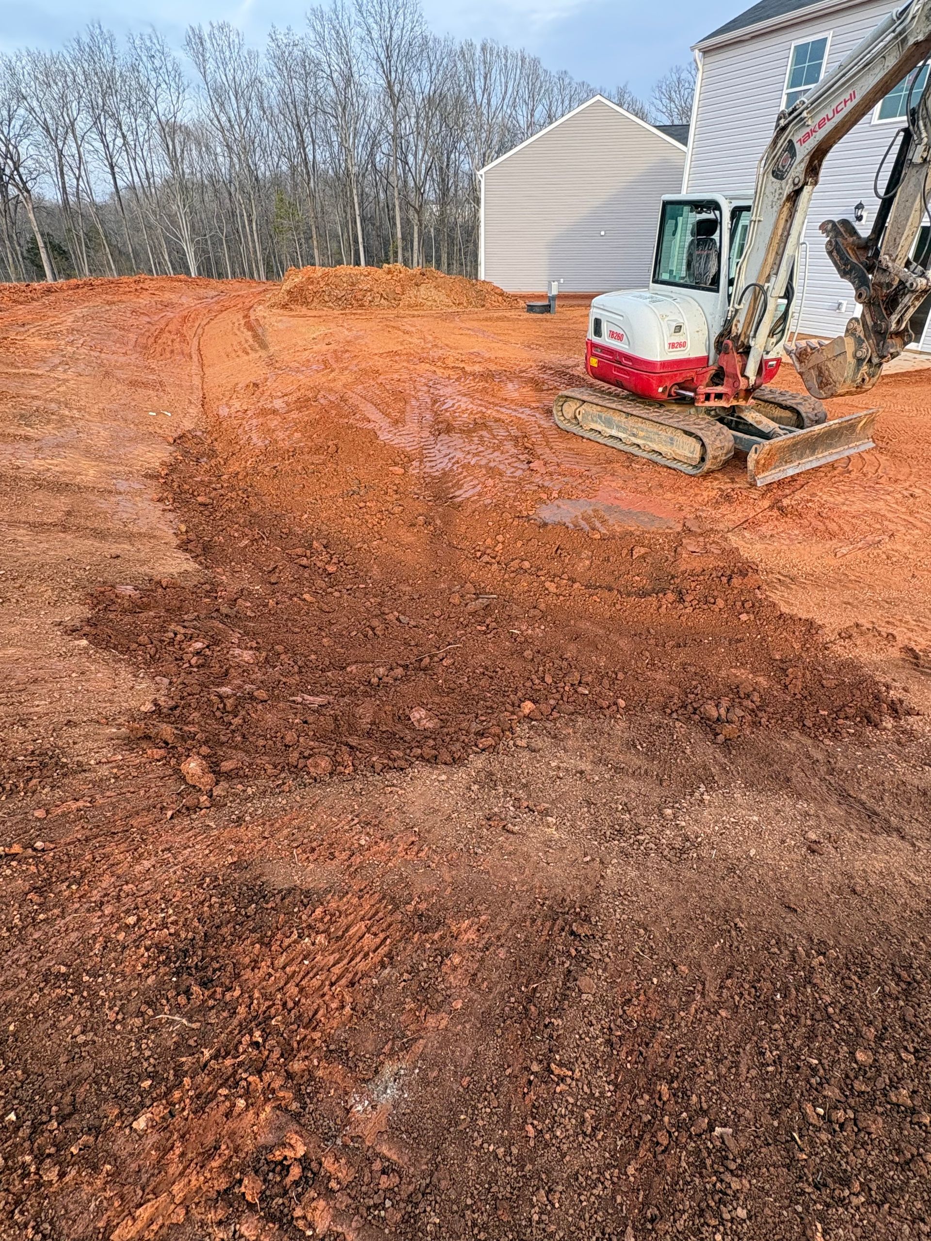 A compact excavator sits on a dirt lot in front of a residential house under construction.