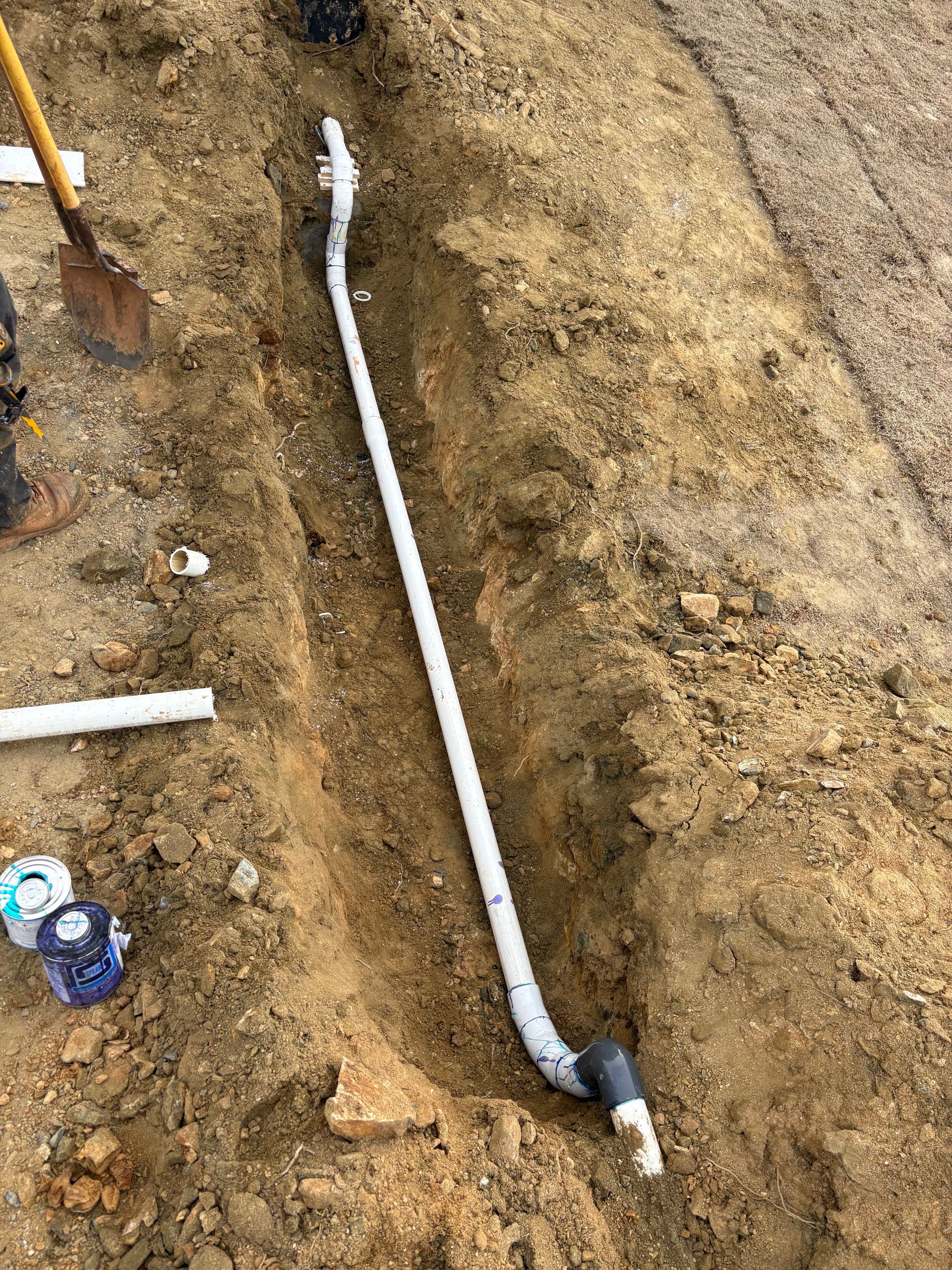 A long PVC pipe lies in a narrow, freshly dug dirt trench, with a shovel nearby and construction supplies in the corner.