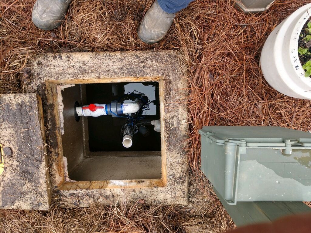 Open septic tank with pipes, a valve, and standing water. Person's boots are visible.