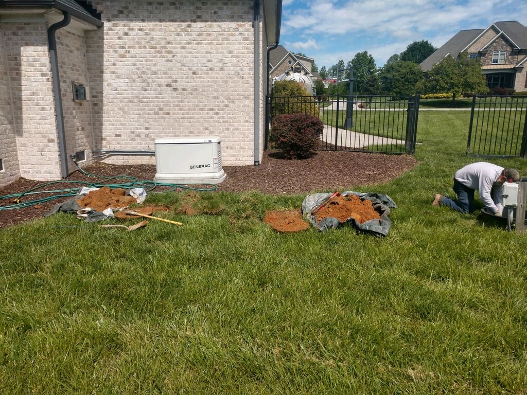 Person kneeling in yard near dirt piles and a generator. House and fence in background.