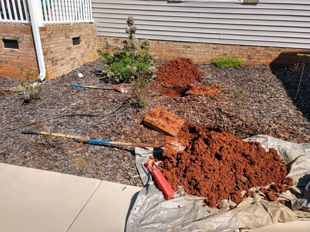 A front yard flowerbed with dug-up red clay, tools, and mulch, next to a house with siding.