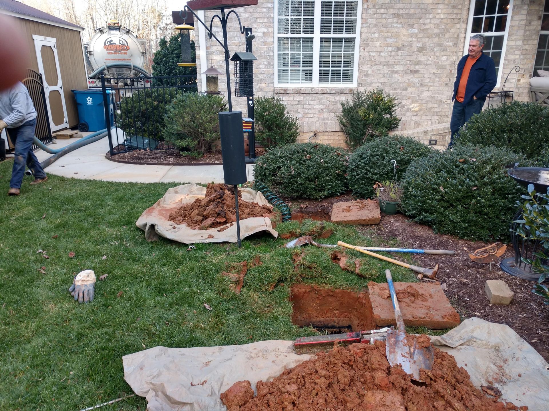 Yard with dirt pile, bricks, and workers; brick house in background.