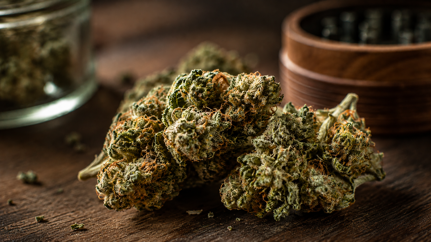 Close-up of cannabis flower buds on a rustic wooden surface with a glass jar and wooden grinder in t