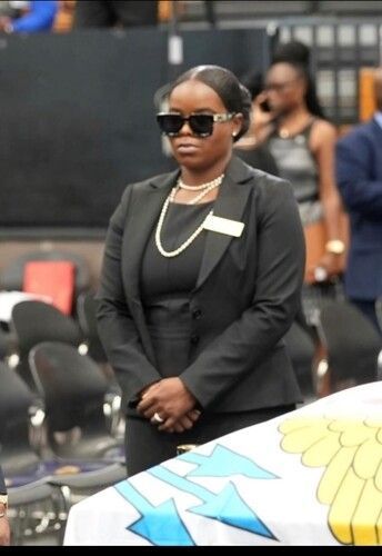 Woman in black suit and sunglasses standing at an outdoor event, with chairs and people in the background