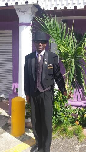 Person in a black suit and hat stands outside a purple building beside a yellow bollard.