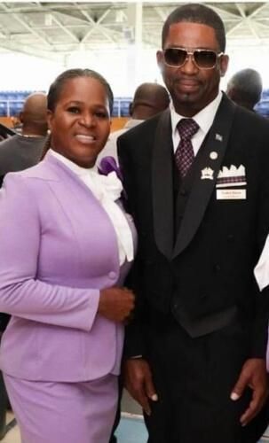 Two formally dressed people posing together indoors, one in a lavender suit and one in a black suit with medals.