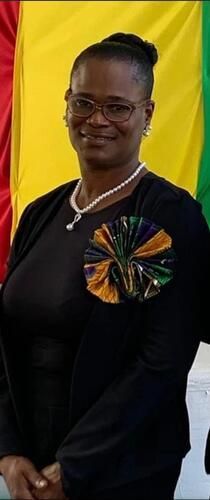 Portrait of a person in black attire with a colorful ribbon pin, standing before yellow and green drapes