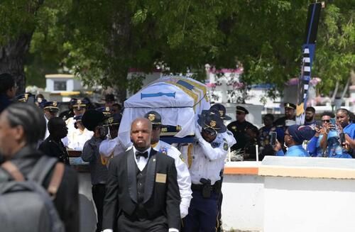 Mourners carry a white casket in a funeral procession outdoors, with a suited man leading in front.