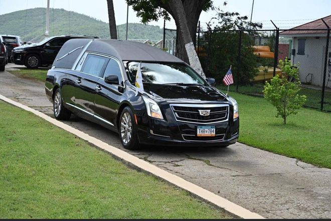 Black hearse parked on a driveway beside a grassy lawn, with a house and hills in the background