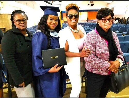 Four people posing indoors, one in a graduation cap and gown holding a diploma folder.