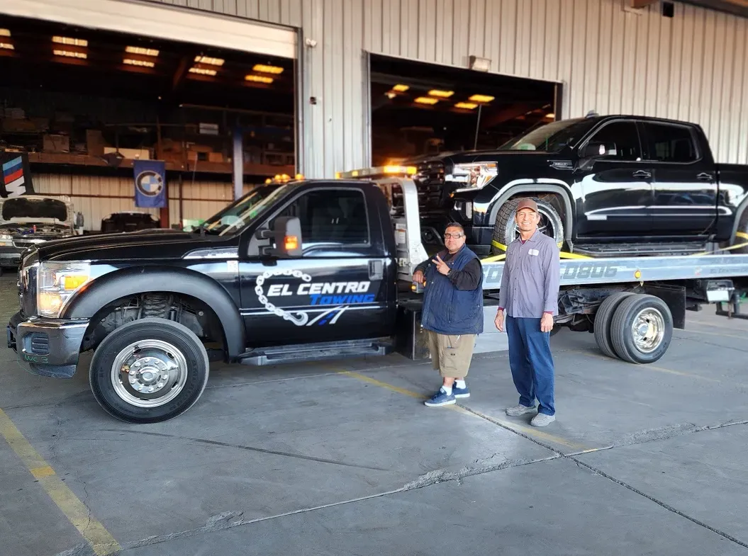 Two men are standing in front of a tow truck with a black truck on the back.