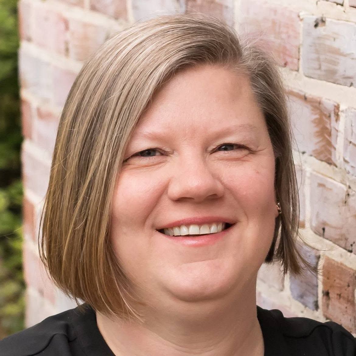 Smiling person with short blond hair in a black top, standing in front of a brick wall