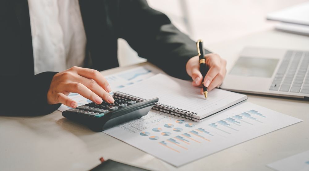 Hands using calculator and pen over financial charts beside a laptop on a desk
