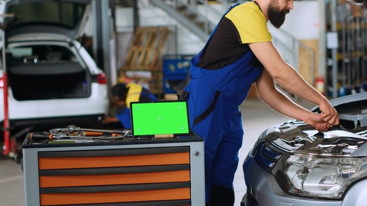Mechanic in blue overalls working on a silver car in a garage, with a toolbox nearby