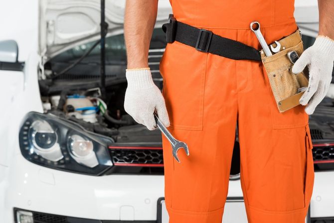 Mechanic in orange coveralls holding tools beside a white car with the hood open