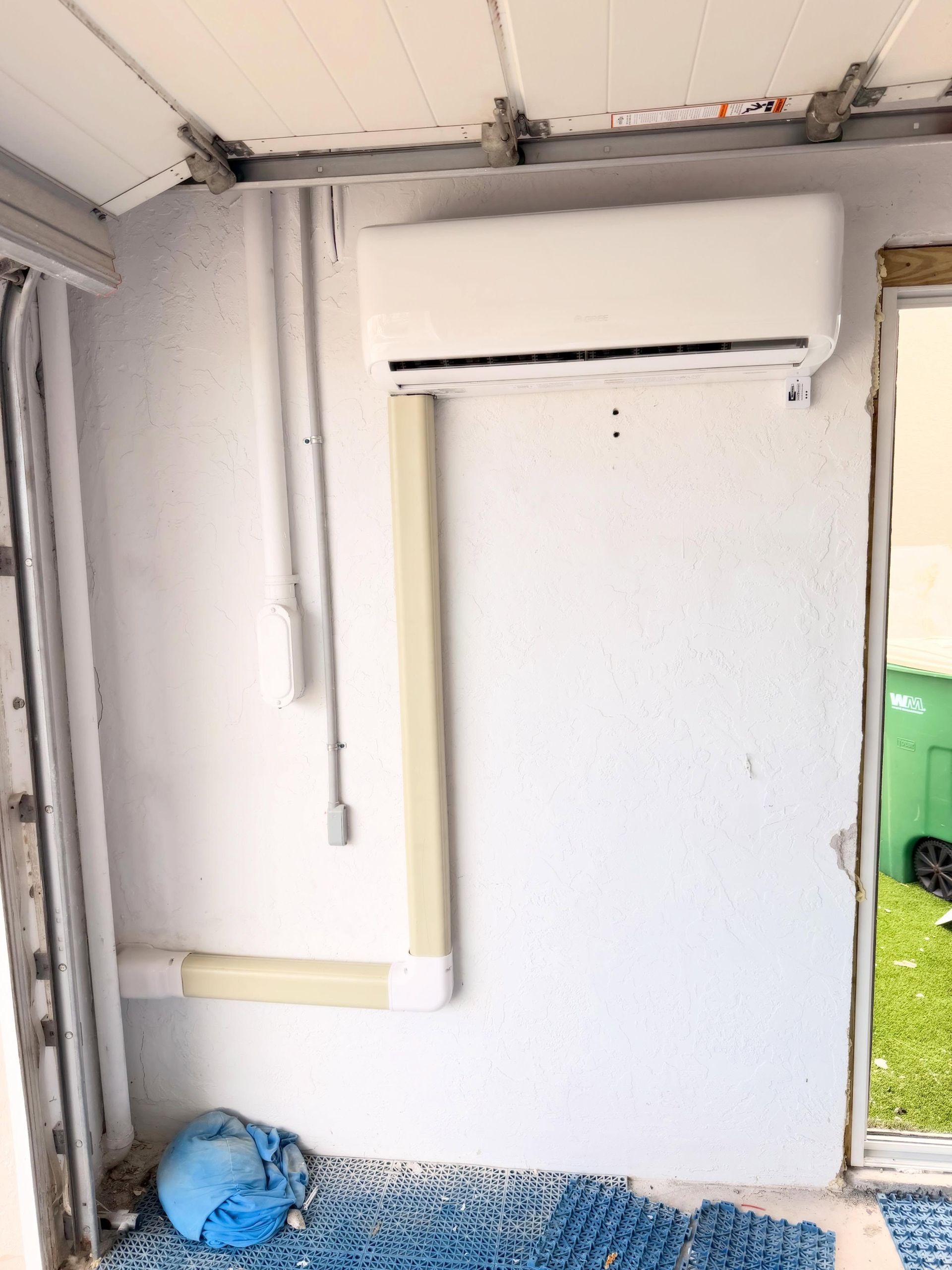 Empty white utility room with exposed pipes and wiring, blue bag on the floor, and an open doorway to the right
