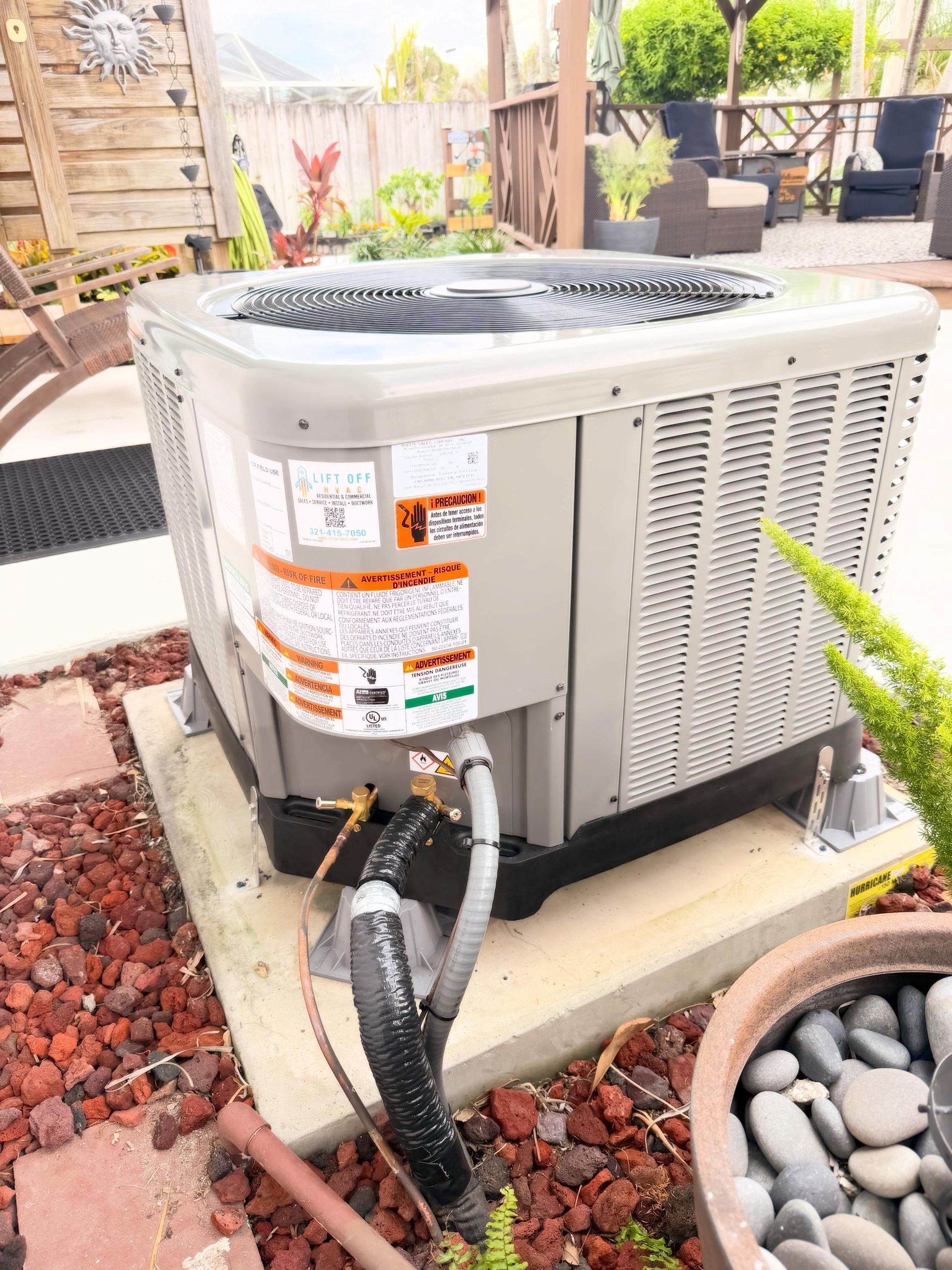 Outdoor air conditioning unit beside a patio, with black hoses and red gravel around the base.