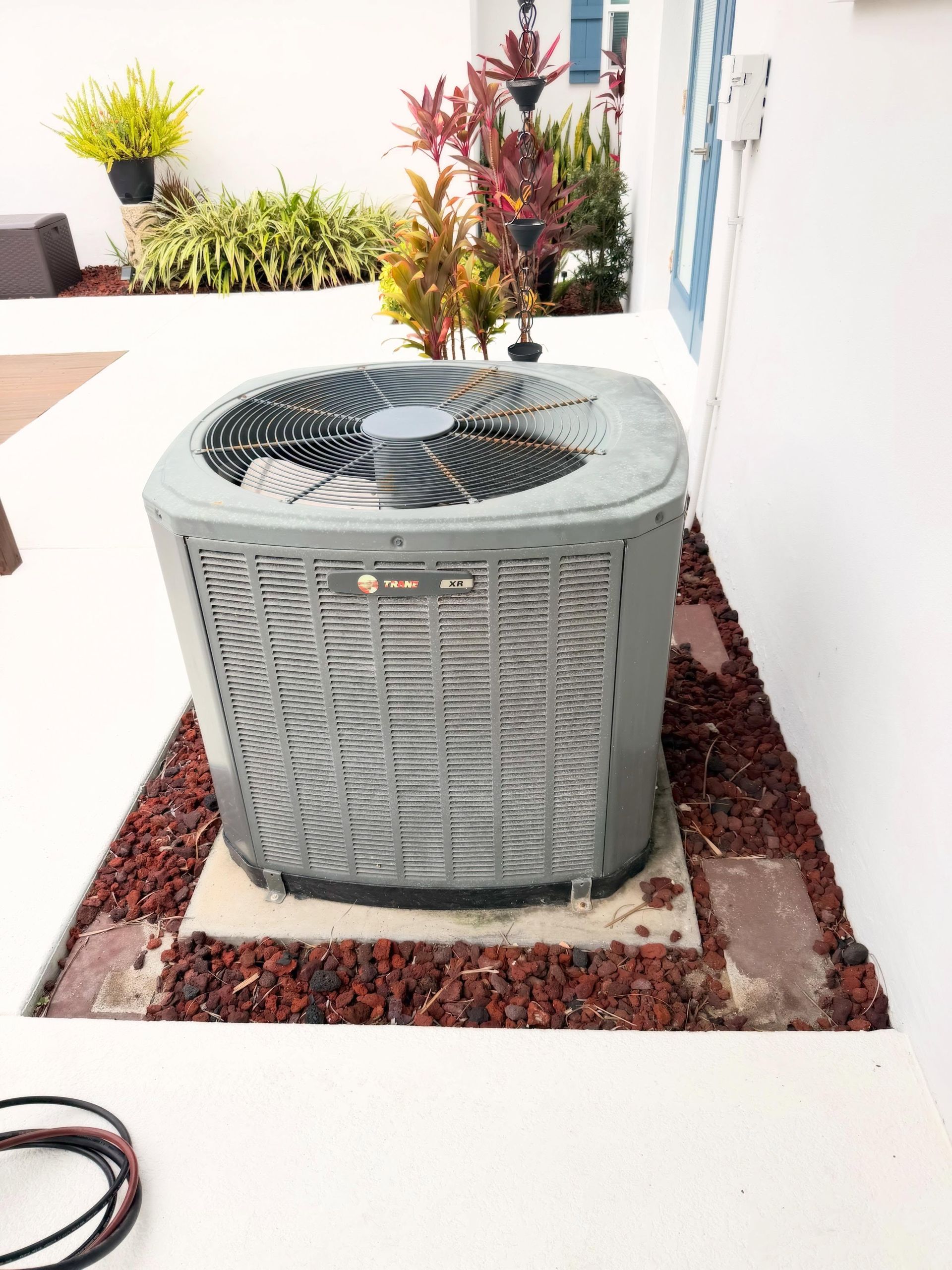 Outdoor HVAC unit beside a white wall, bordered by red mulch and desert plants on a patio.