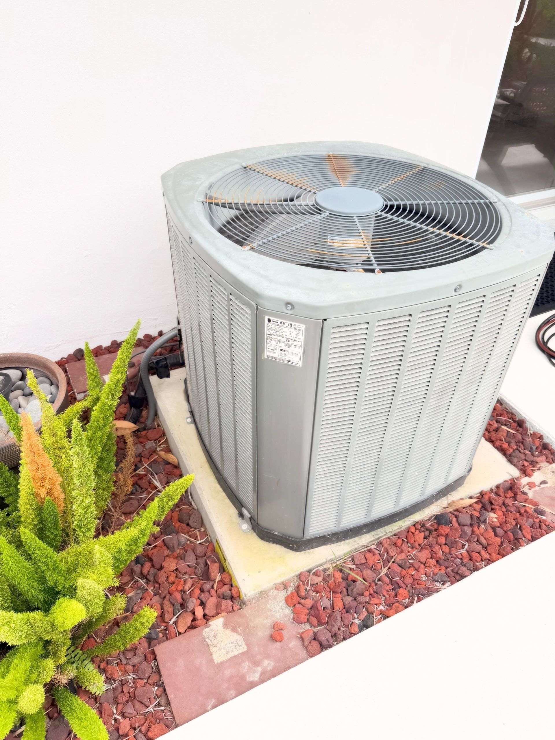 Outdoor air conditioning unit beside a white wall, bordered by red rocks and green plants.