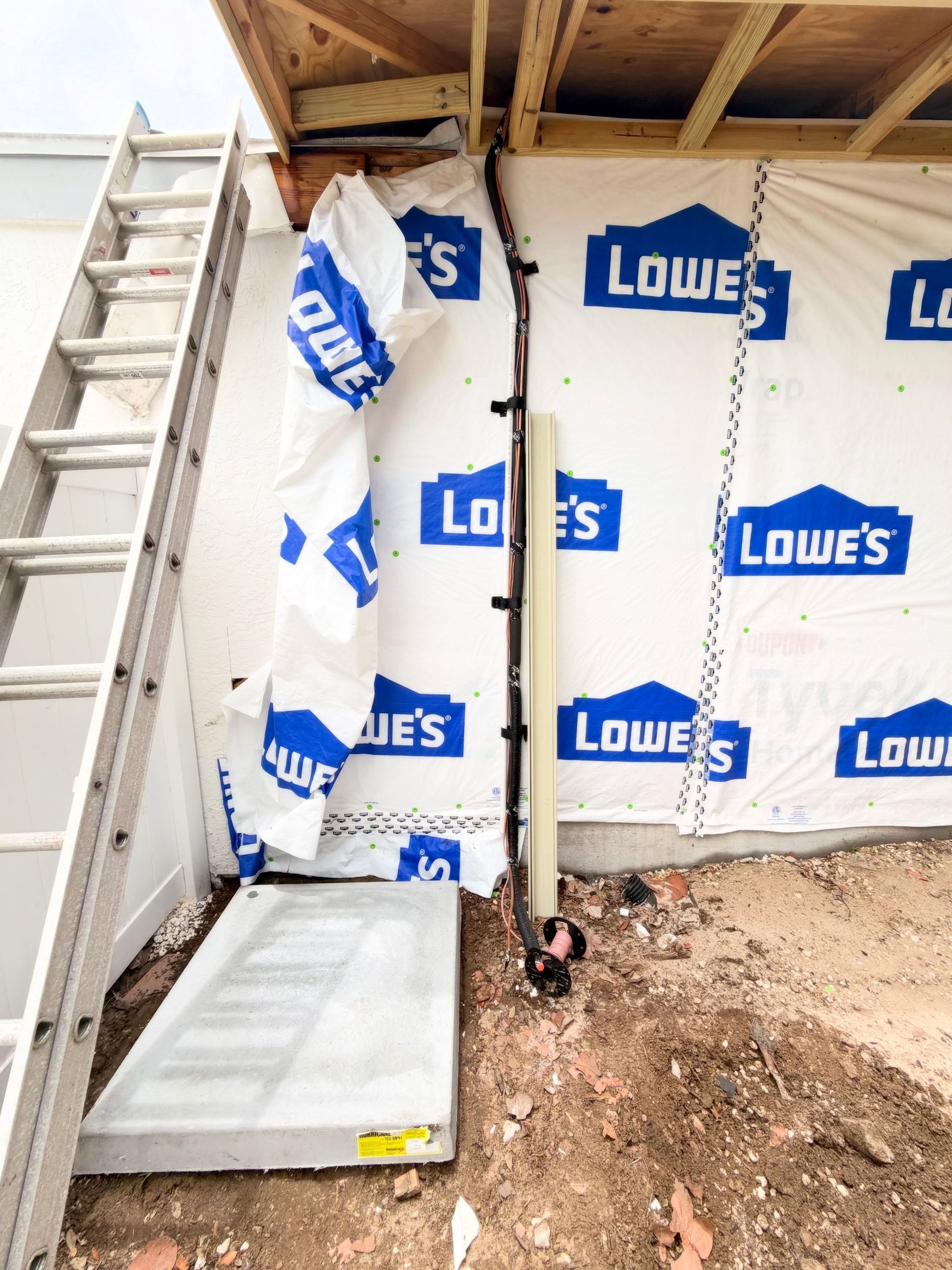 Ladder beside house wall wrapped in Lowe's house wrap, with exposed dirt and construction debris