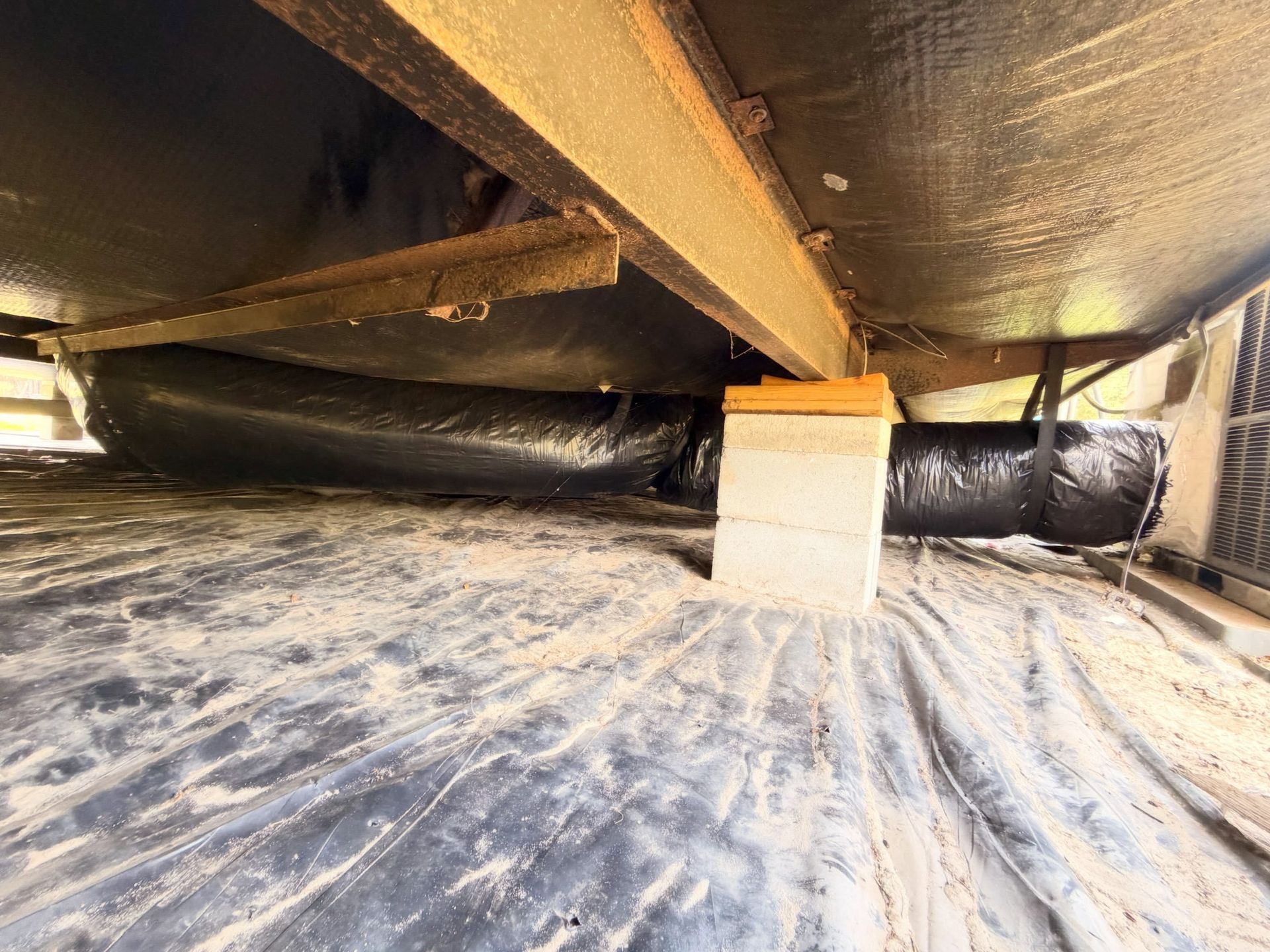 Dusty crawl space with black insulated pipes and a wooden support beam under a floor