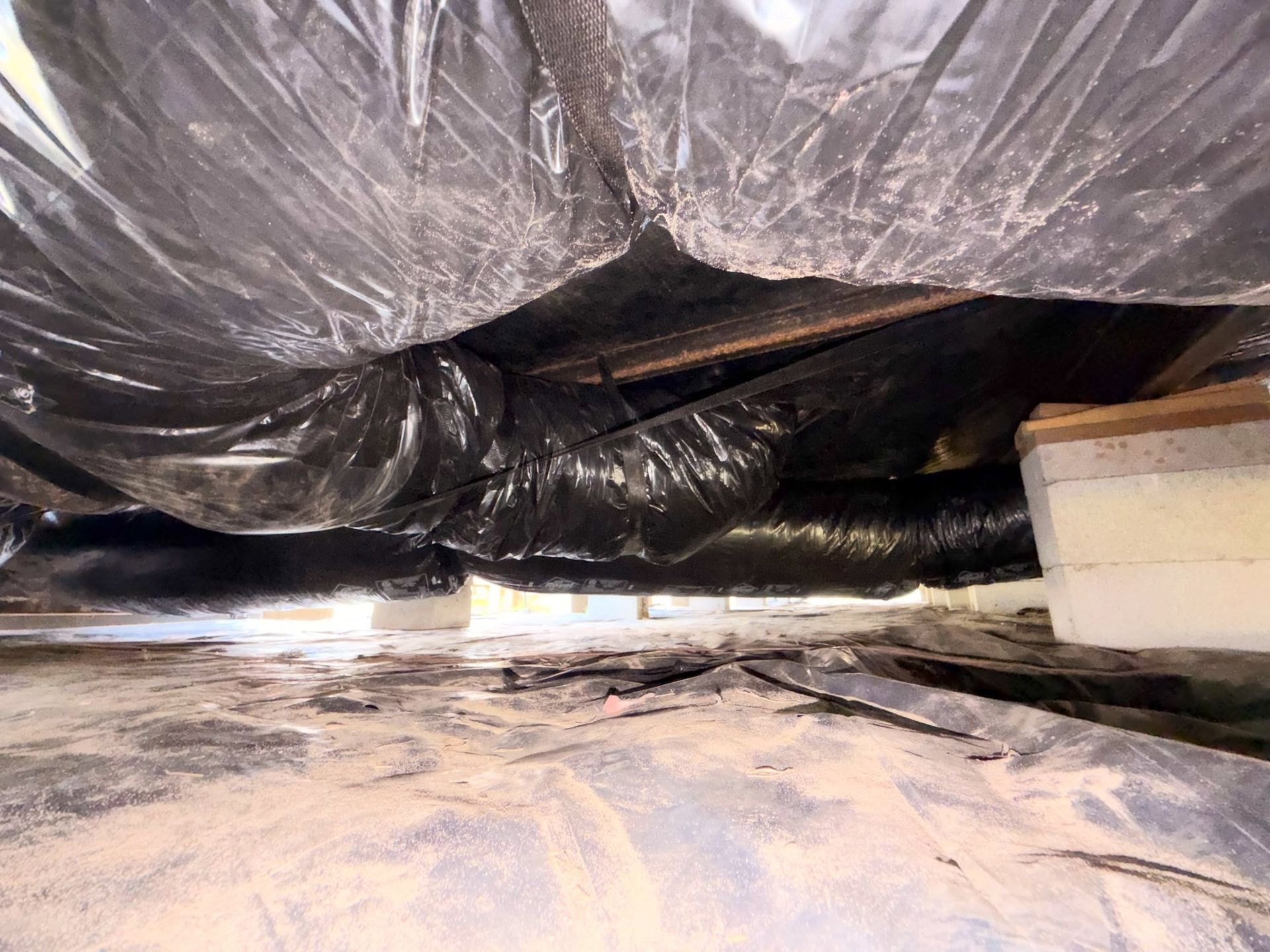 Plastic sheeting draped over a crawlspace with debris on the floor and a white box at right