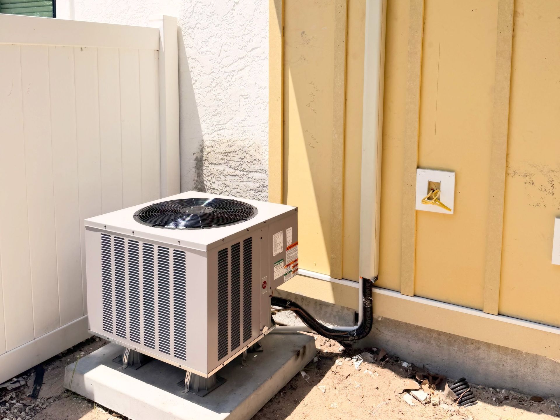 Outdoor air conditioner unit beside a beige wall and white fence in a narrow yard