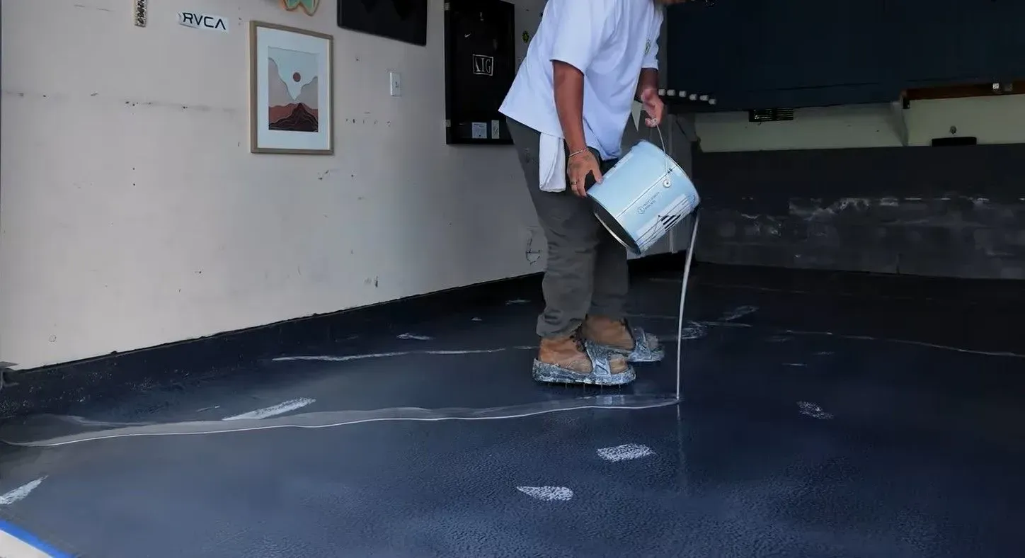 Person pouring liquid onto a dark garage floor from a metal bucket