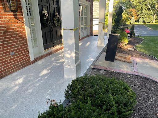 A freshly coated, speckled gray porch leads to a brick building with a black front door. A green bush is in the foreground.