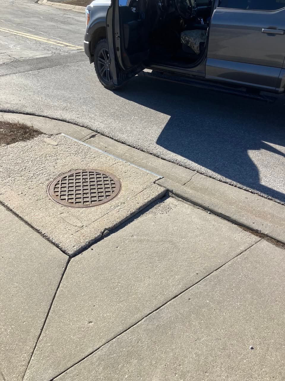 A dark-colored pickup truck with its door open, parked next to a curb with a manhole cover.