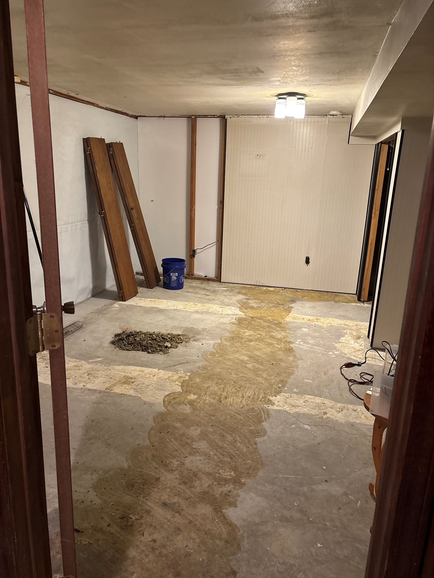 Basement room with unfinished walls, concrete floor, wooden boards, and a blue bucket.