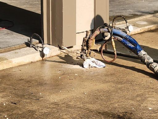 A rusty fuel pump nozzle and hoses on a concrete ground, beside a beige pillar.