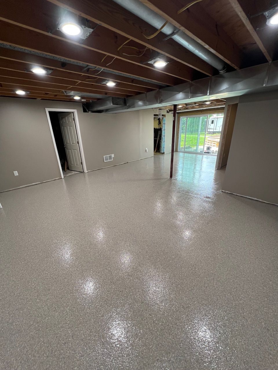 Empty unfinished basement with gray floor, exposed ceiling beams, and sliding glass door to backyard.