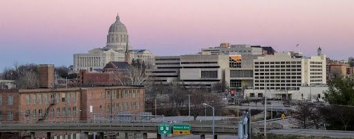 City skyline at sunset with the State Capitol dome visible on the horizon; featuring several buildings and highway signs.