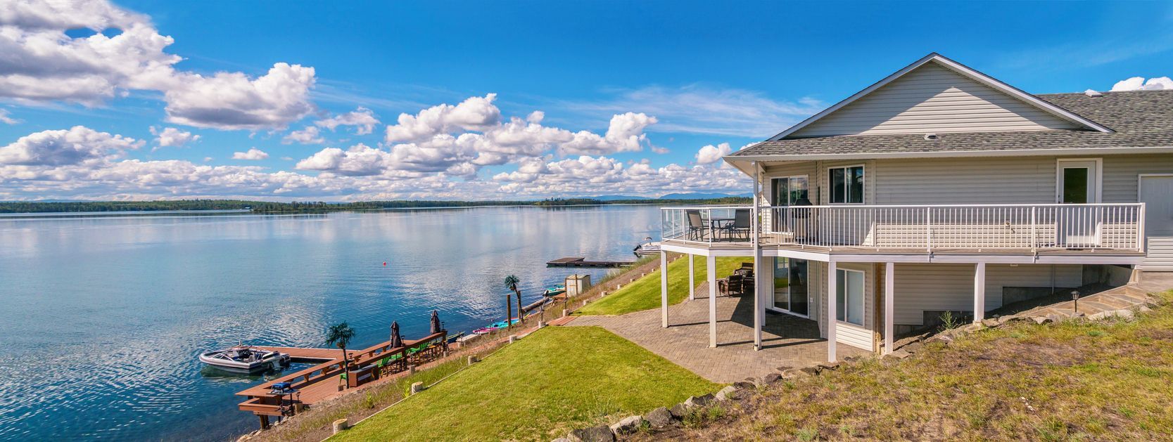 A lakeside house with a deck, docked boats, and a bright blue sky with fluffy clouds.