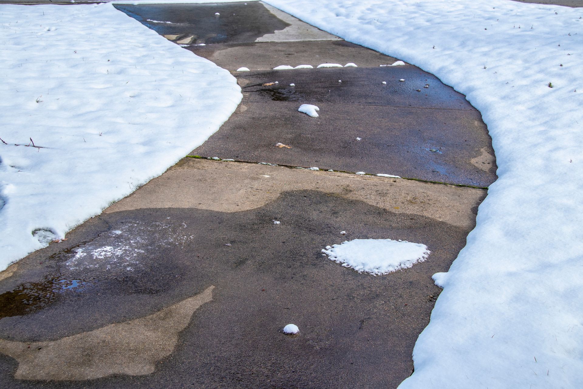 Snow-covered curved sidewalk with patches of melting snow. The pavement is dark gray, with white snow on either side.