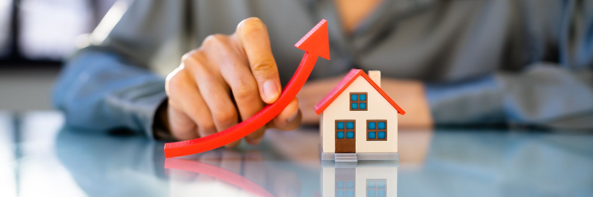 A hand holds a red arrow pointing upwards over a toy house, representing rising real estate value.