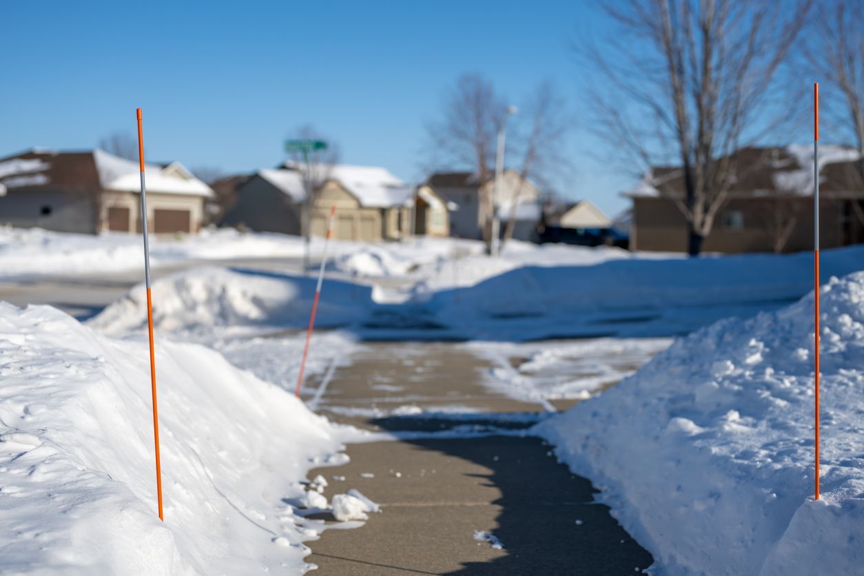 Snowy suburban sidewalk with orange snow markers, plowed path, and houses under a clear blue sky