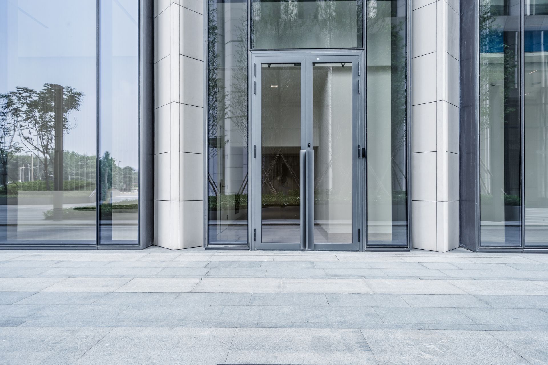 Glass entrance doors to a modern building, with light gray stone paving leading up to them.